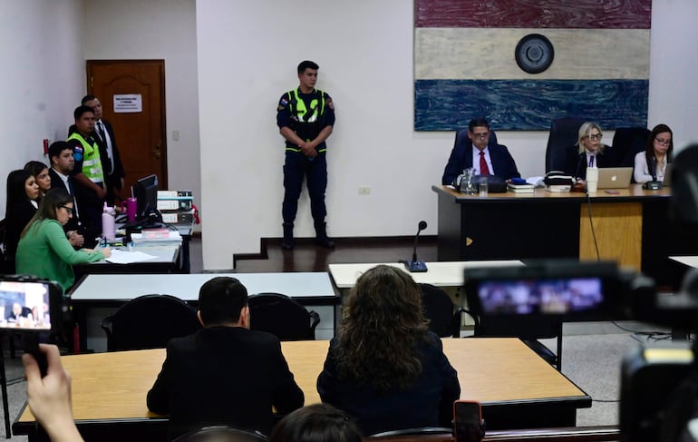 Mujer toma notas mientras un hombre con traje oscuro habla; seguridad en uniforme observa en un ambiente formal y decorado con la bandera paraguaya.