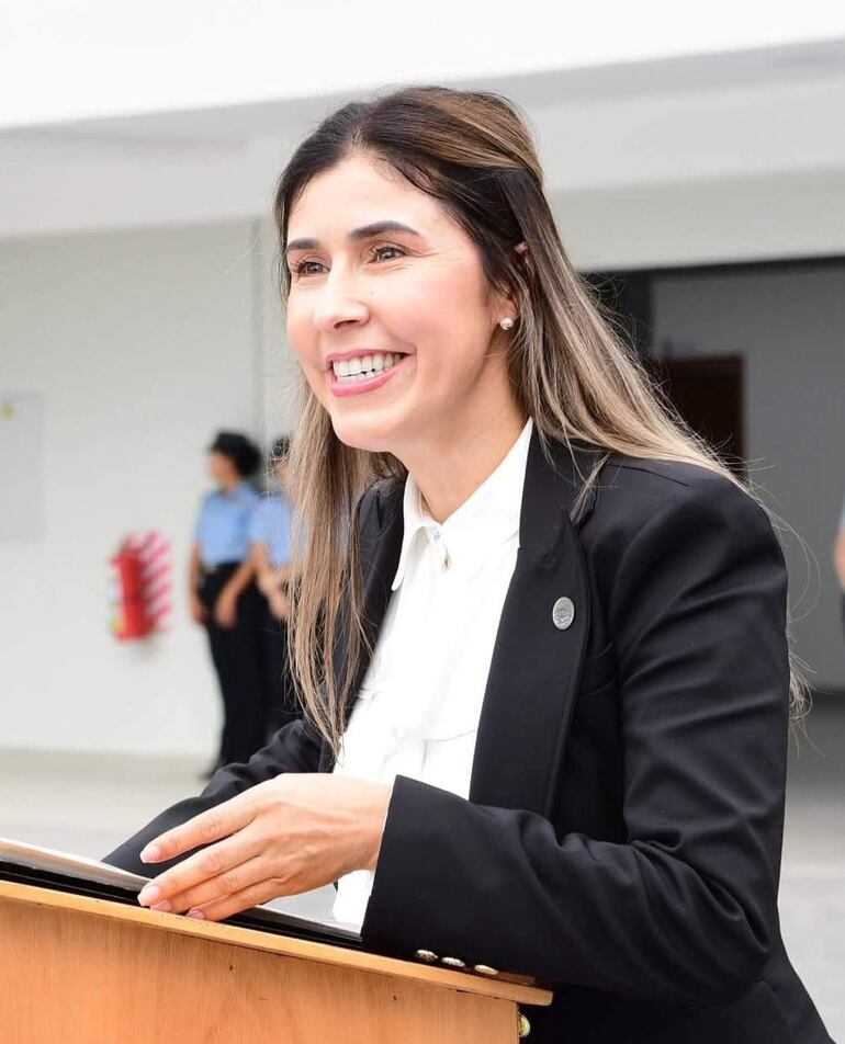 Mujer sonriente en podio con blazer negro y blusa blanca, rodeada de asistentes en un ambiente formal.