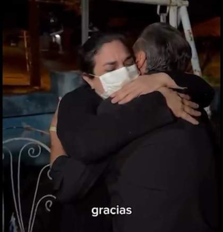 Dos personas con ropa oscura se abrazan. Una lleva mascarilla y parece conmocionada, mientras la otra ofrece consuelo.