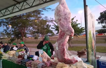 Hombre con gorro verde y delantal sostiene un teléfono, rodeado de productos alimenticios en un ambiente festivo.