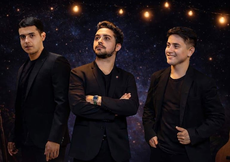 Tres hombres en chaquetas y camisetas negras, el central con brazos cruzados, rodeado de una atmósfera festiva iluminada.