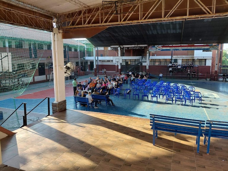 Grupo de personas sentadas en sillas azules, atentas a un orador en ambiente escolar. Espacios deportivos al fondo.