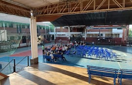 Grupo de personas sentadas en sillas azules, atentas a un orador en ambiente escolar. Espacios deportivos al fondo.