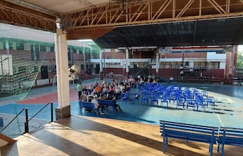 Grupo de personas sentadas en sillas azules, atentas a un orador en ambiente escolar. Espacios deportivos al fondo.
