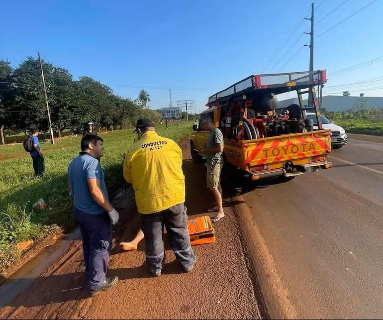 Hombre en camiseta azul atiende a persona en el suelo, rodeados de vegetación y un vehículo Toyota amarillo al fondo.