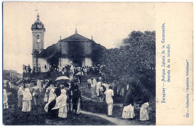Multitud de personas con atuendos tradicionales y paraguas, reunidas frente a la Antigua Iglesia de la Encarnación con su imponente arquitectura.