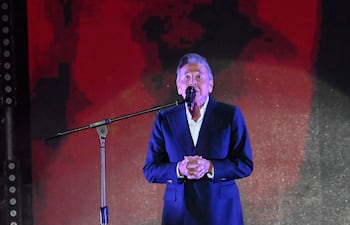 Hombre de mediana edad en traje oscuro y camisa blanca habla frente a un micrófono en escenario de fondo rojo.