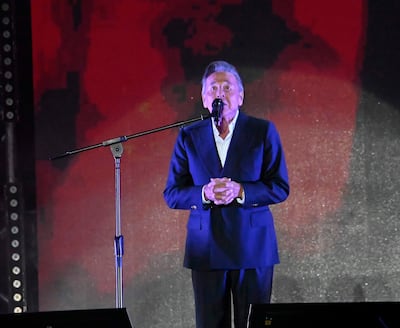 Hombre de mediana edad en traje oscuro y camisa blanca habla frente a un micrófono en escenario de fondo rojo.