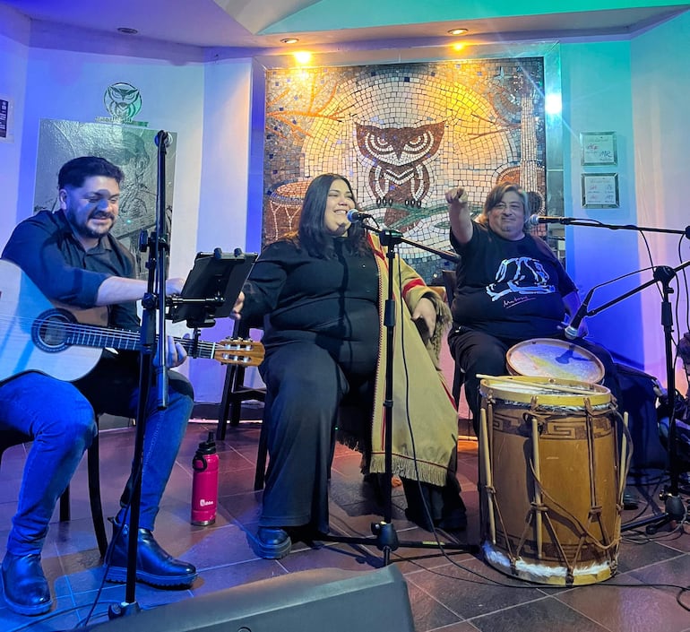 Mujer con micrófono sonriente en atuendo oscuro, acompañada de una guitarrista y otra vocalista en un ambiente decorativo y con luces suaves.