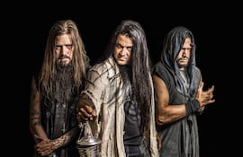Tres hombres de cabello largo y barba en un fondo oscuro, el del centro sostiene un objeto metálico, mientras los otros lo miran atentos.