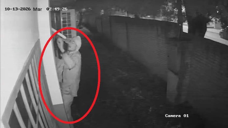 Persona en prenda clara intentando acceder a través de una ventana, en un ambiente iluminado tenuemente.