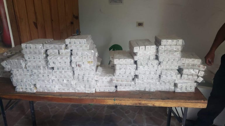 Cajas de cigarrillos apiladas en mesa de madera, con un vigilante de pie a la derecha en un ambiente interior claro.