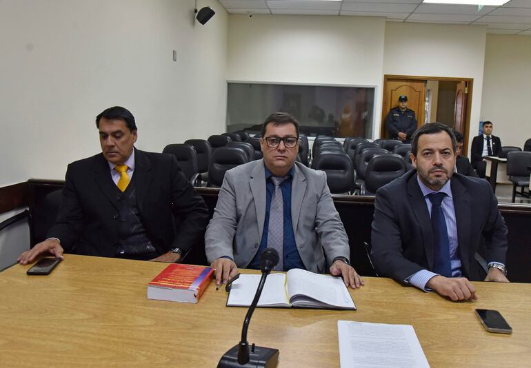 Tres hombres formales sentados en mesa de tribunal, micrófonos y un libro frente a ellos, en ambiente sobrio y formal.