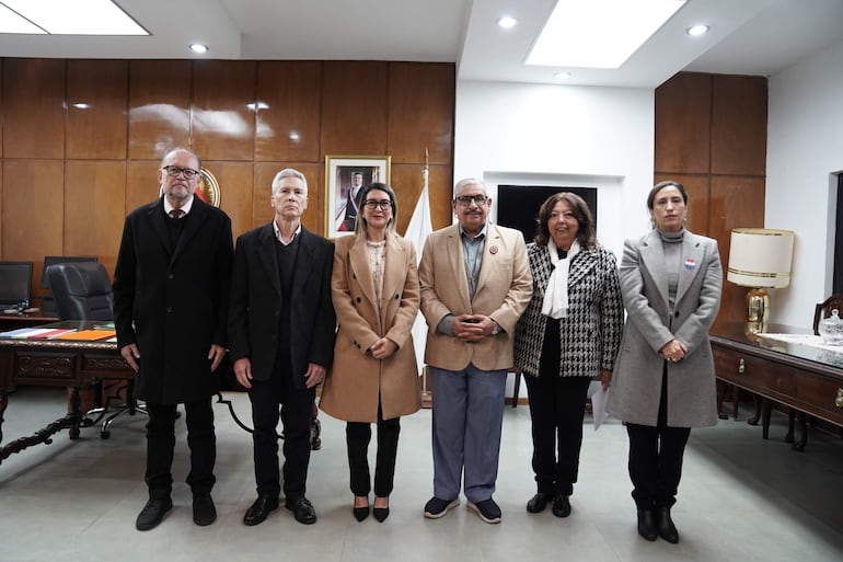 Mirtha Arias y cinco consejeros sonríen, todos en trajes formales, en una sala con fondo de madera y lámpara de escritorio.