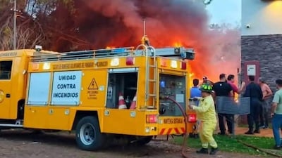 Bombero vestido con uniforme amarillo opera una manguera mientras testigos observan el incendio de cubiertas, generando humo negro.