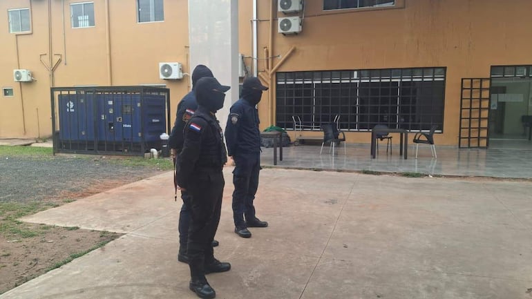 Tres hombres en uniformes oscuros y pasamontañas, en un ambiente exterior con un edificio naranja al fondo.