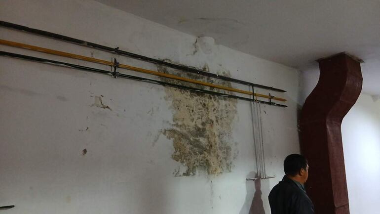 Hombre de espaldas observa la pared deteriorada con manchas de humedad y tubos expuestos en el hospital.