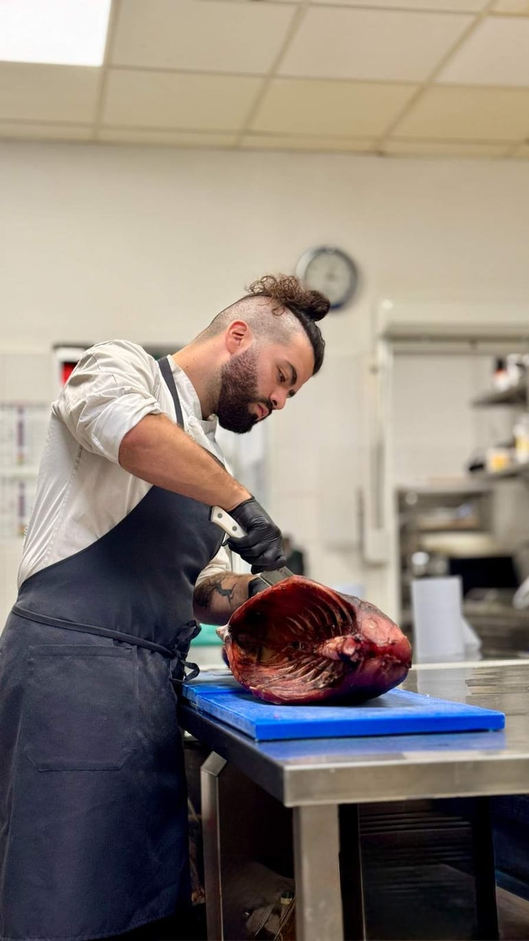 Alejandro Calcaterra, hombre con barba y camisa blanca, corta pescado en una tabla azul, con utensilios de cocina en el fondo.