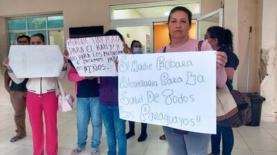Mujer con suéter rosa y expresión seria sosteniendo cartel sobre salud, rodeada de manifestantes en edificio público.