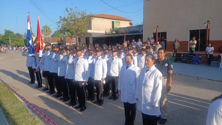 Grupo de estudiantes en uniformes blancos saludan con banderas, rodeados por una audiencia atenta en un día despejado.