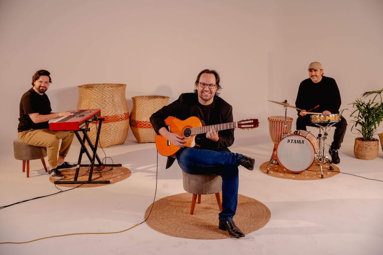 Diego Guzmán sonríe tocando la guitarra, mientras un tecladista y un baterista lo observan en un estudio acogedor.