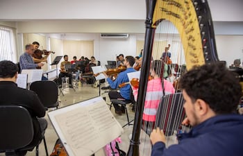 Músicos de la Orquesta Sinfónica del Congreso Nacional ensayan, con director mayor de gafas al frente y varios instrumentos visibles.