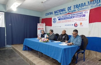 Cuatro hombres en mesa con mantel azul, algunos escribiendo y otros escuchando, en salón decorado con bandera paraguaya.