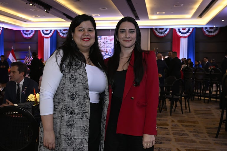 Mujeres sonrientes en evento formal, una con chaleco blanco y negro, otra con blazer rojo, rodeadas de decoración festiva.