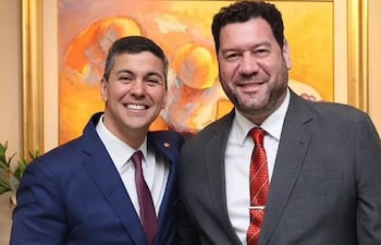 Peña en traje azul y corbata burdeos, Cappello en traje gris y corbata roja, ambos sonriendo en una sala decorada elegantemente.