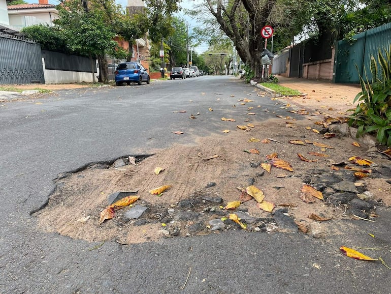 Calle en mal estado con baches y hojas secas, rodeada de casas y vehículos estacionados, sin personas visibles.