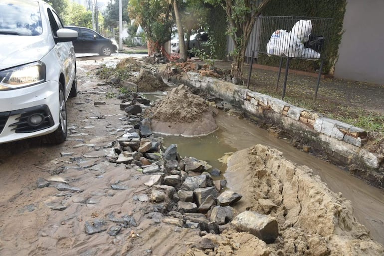 Calle en mal estado con lodo y piedras sueltas, dos vehículos visibles y acumulación de agua a un lado.