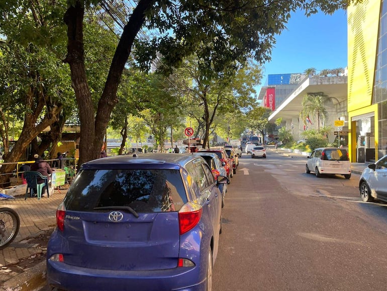 Personas sentadas y de pie en una calle soleada, rodeados de árboles y vehículos estacionados, frente a un edificio con cartel 'Ameris'.