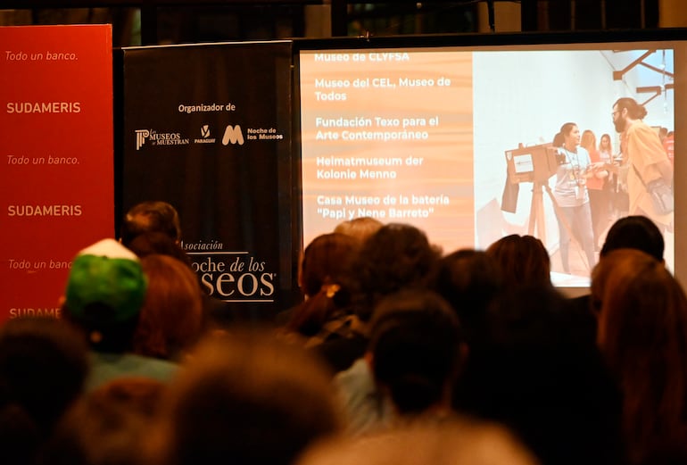 Multitud de personas de espaldas, escuchando atentamente una presentación sobre museos en un ambiente cerrado.