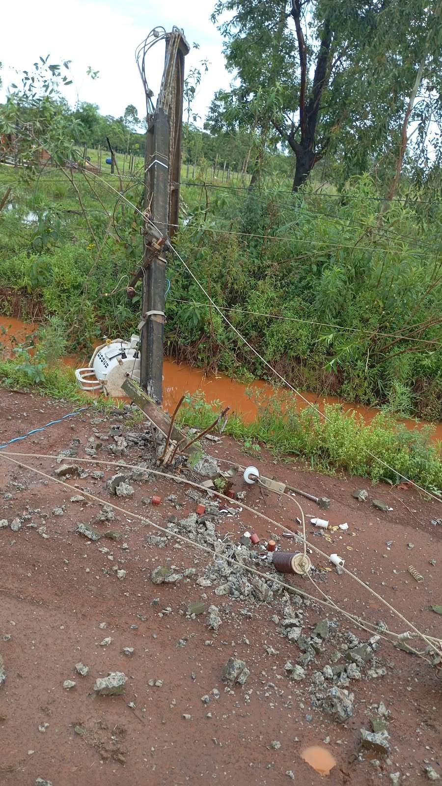 Poste de electricidad inclinado hacia un lado, rodeado de escombros en un paisaje rural con vegetación densa.