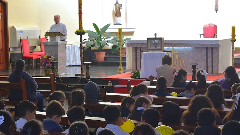 Sacerdote canoso en altar hablando a grupo de niños en bancos, ambiente solemne con velas y decoración religiosa.