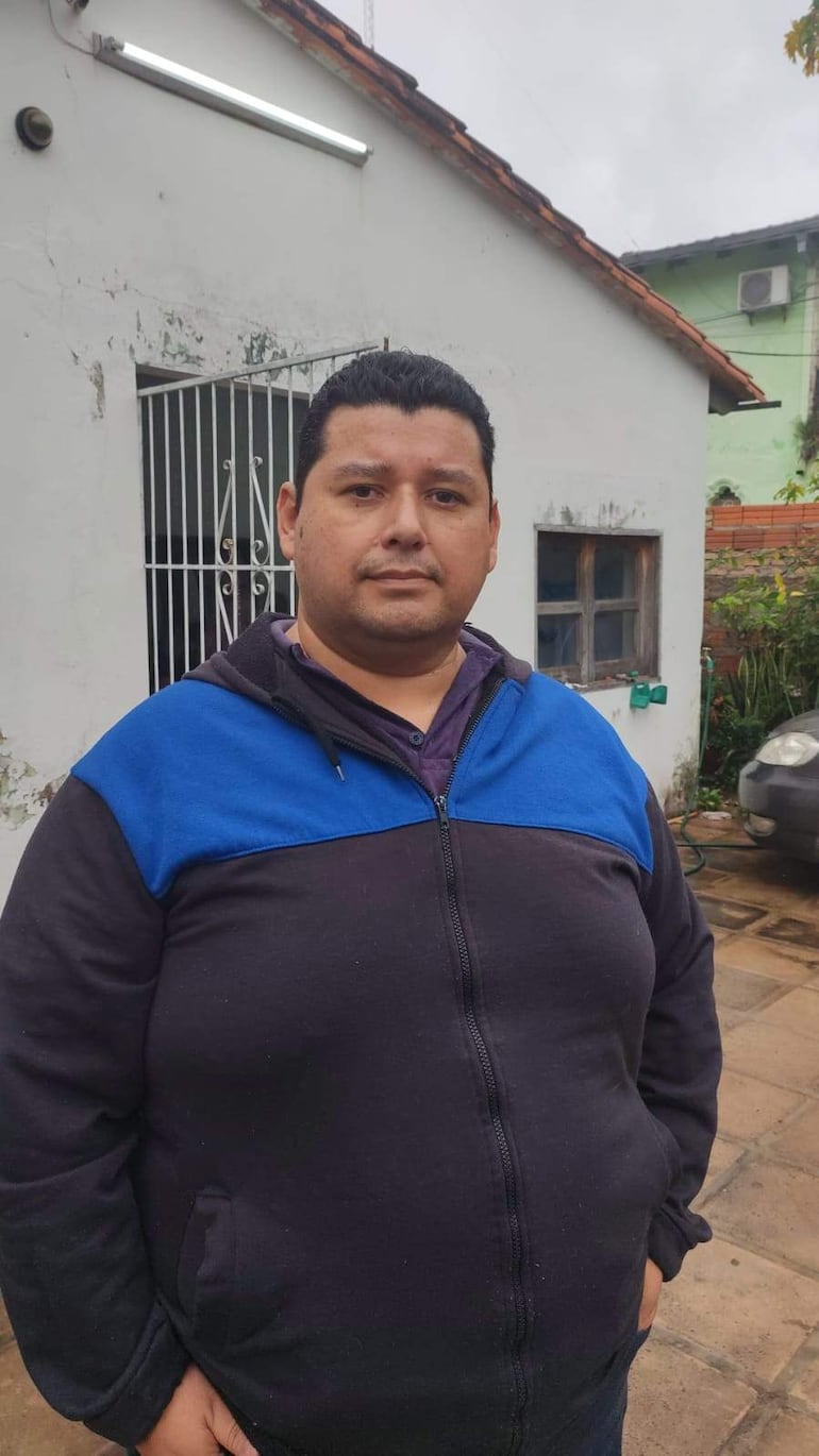 Hombre robusto de pie con chaqueta oscura y azul frente a una vivienda, sin otras personas presentes.
