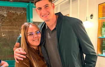Mujer de cabello largo y chaqueta marrón sonríe junto a hombre alto con chaqueta verde en un café con paredes de ladrillo.