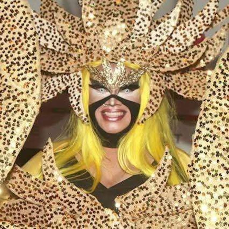 Persona con disfraz dorado y máscara grande, cabello amarillo, en un ambiente festivo.