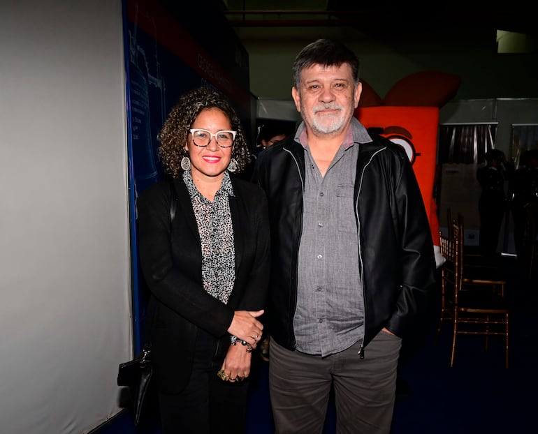 Mujer con blusa de estampado animal y anteojos, junto a hombre de cabello canoso y chaqueta negra, sonriendo en un evento formal.