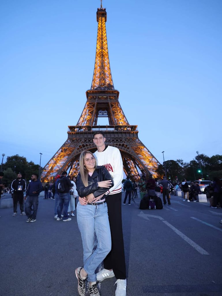 Pareja abrazándose, ella con chaqueta negra y jeans claros, él con camiseta blanca y pantalones oscuros, Torre Eiffel iluminada de fondo.