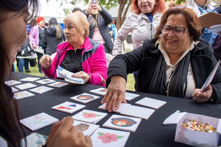 Tres mujeres concentradas en juego de cartas, con chaquetas en colores negro y fucsia, rodeadas de un ambiente festivo al aire libre.