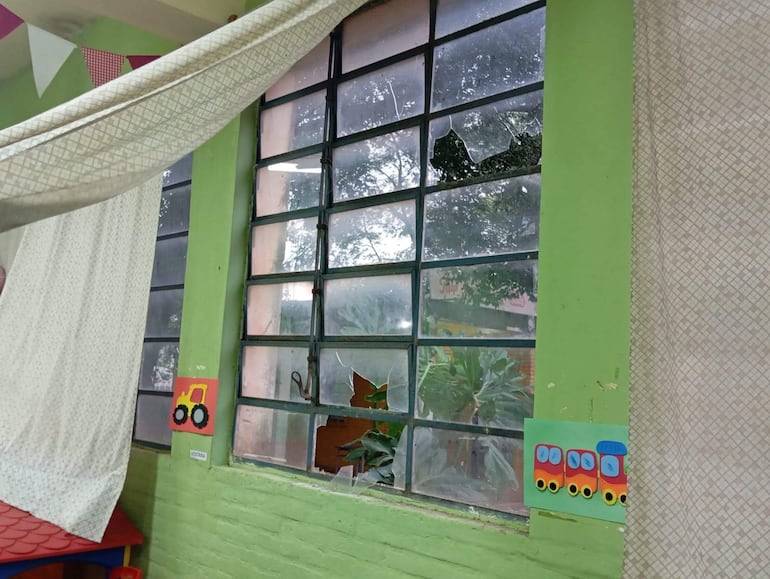 Interior de salón de clases con pared verde, ventana rota y dibujos infantiles en las paredes.