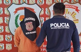 Persona con capucha negra y suéter naranja en posición de detención, acompañada por un policía de camiseta azul con la palabra 'POLICÍA'.