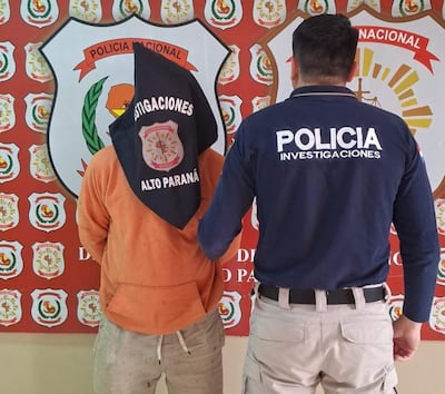 Persona con capucha negra y suéter naranja en posición de detención, acompañada por un policía de camiseta azul con la palabra 'POLICÍA'.