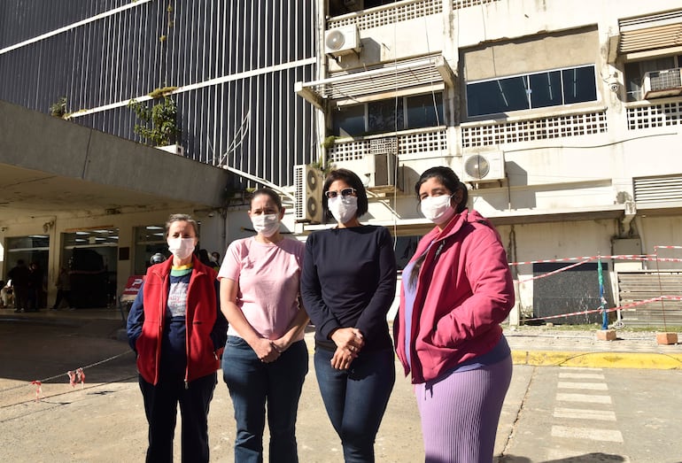 Cuatro mujeres, una con abrigo rojo y otra con chaqueta rosa, conversan al aire libre con mascarillas, frente a un edificio urbano.