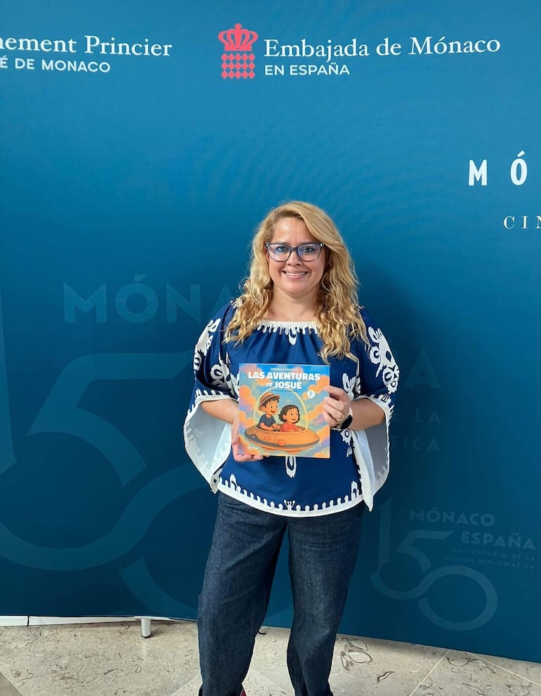 Patricia Franco sostiene su libro 'Las aventuras de Josué' en la Embajada de Mónaco, luciendo blusa azul con blanco.