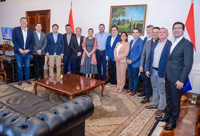 Grupo de hombres y una mujer en vestimenta formal, de pie en una oficina elegante con fondo de pinturas y banderas.