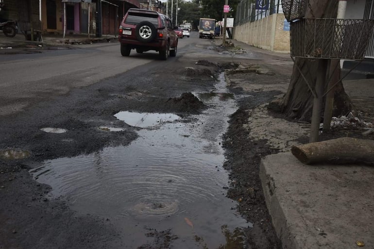 Calle urbana deteriorada con pavimento roto y charcos, dos vehículos en movimiento y comercios cerrados en el fondo.