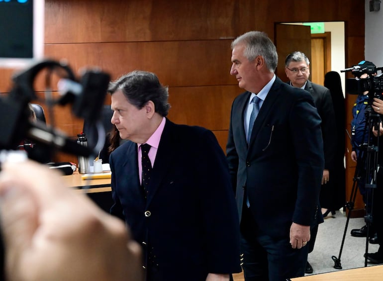 Enrique Fausto Bacchetta Chiriani con chaqueta oscura y camisa rosa, se mueve hacia la salida del salón en un ambiente institucional.