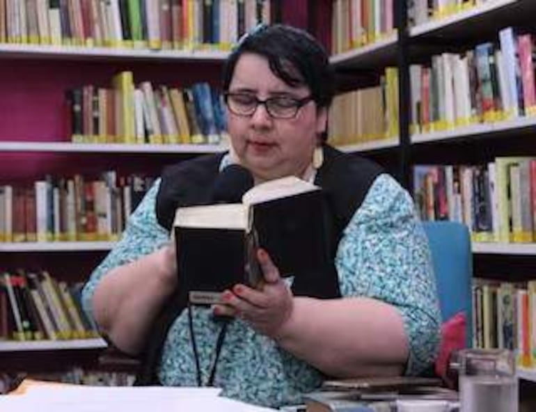 Mujer con gafas y cabello corto, vestida con blusa azul y chaleco negro, leyendo un libro grande en una biblioteca tranquila.